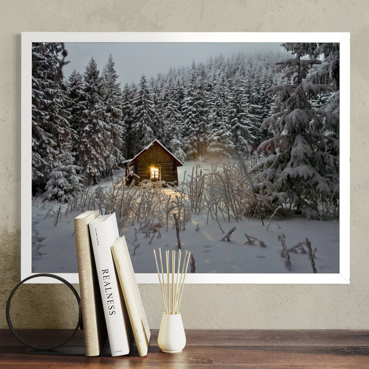 A framed wall art print showing a snowy winter forest with a small cabin and glowing window, displayed above a wooden surface with books and a diffuser against a textured beige wall.