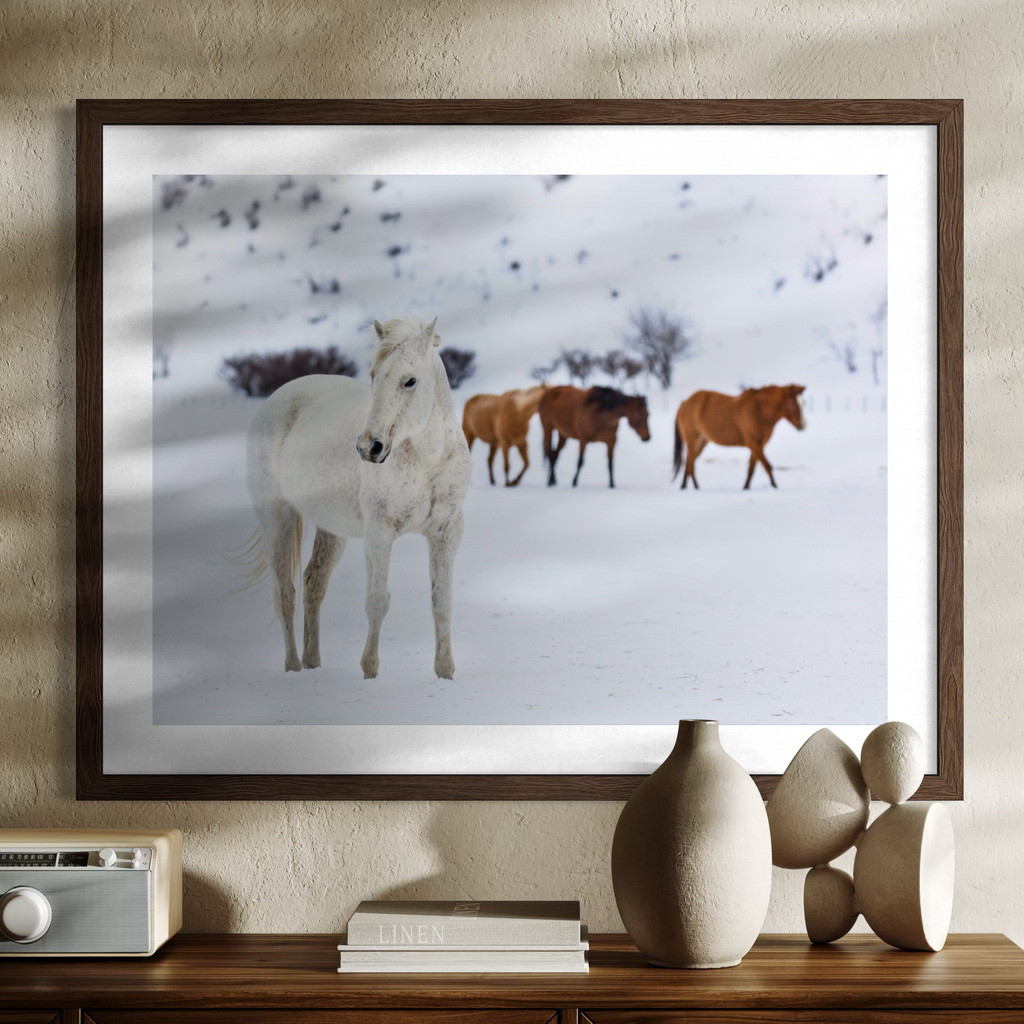 A framed wall art print showing a snowy landscape with a white horse in the foreground and three brown horses behind it, displayed above a console table with decorative vases and books.