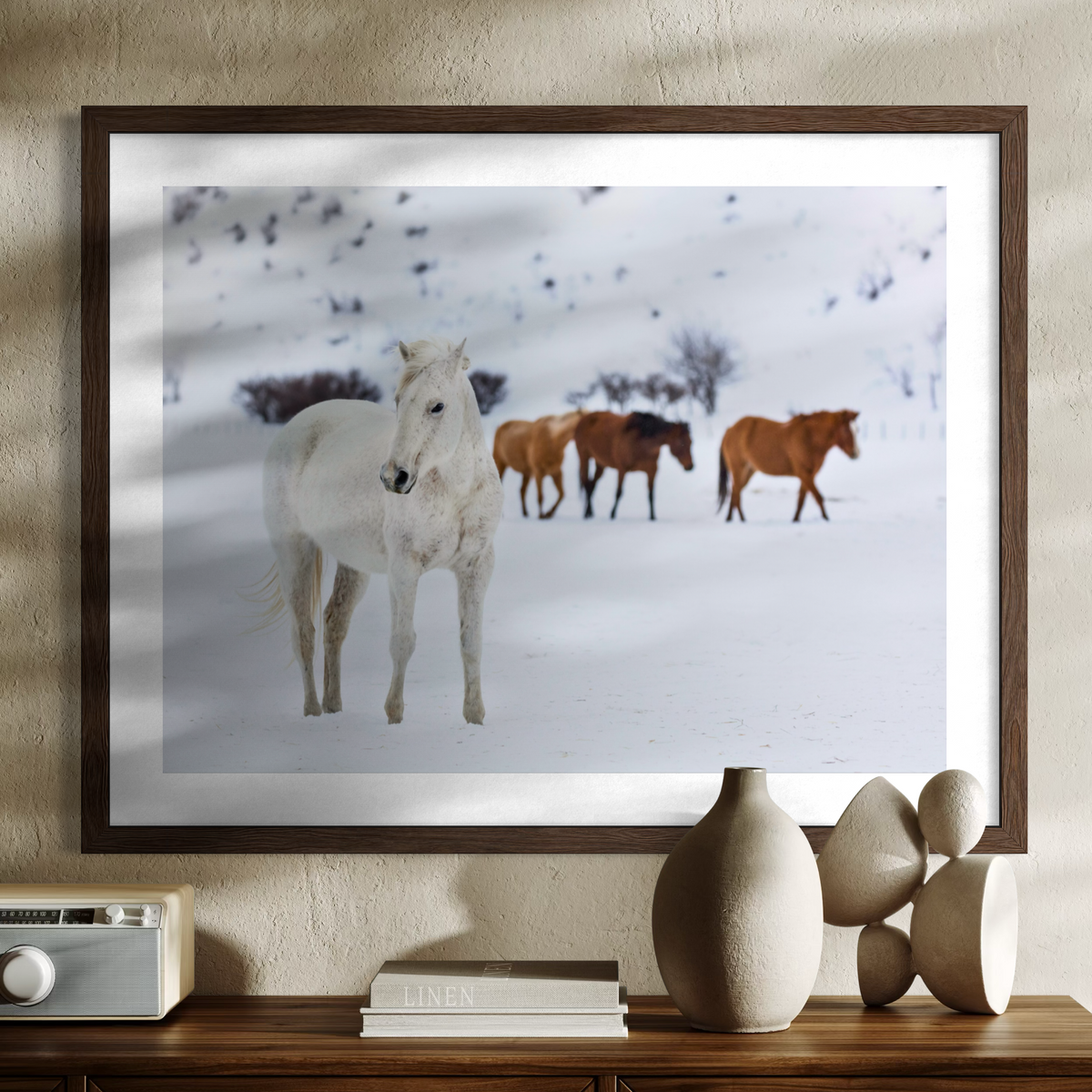 A framed wall art print showing a snowy landscape with a white horse in the foreground and three brown horses behind it, displayed above a console table with decorative vases and books.