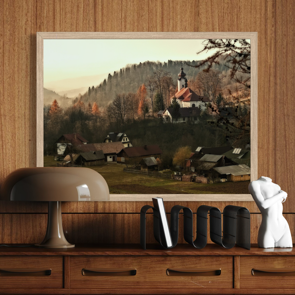 A framed landscape art print depicting a quaint village with a church on a hill, displayed on a wooden shelf next to a lamp and a white bust sculpture.