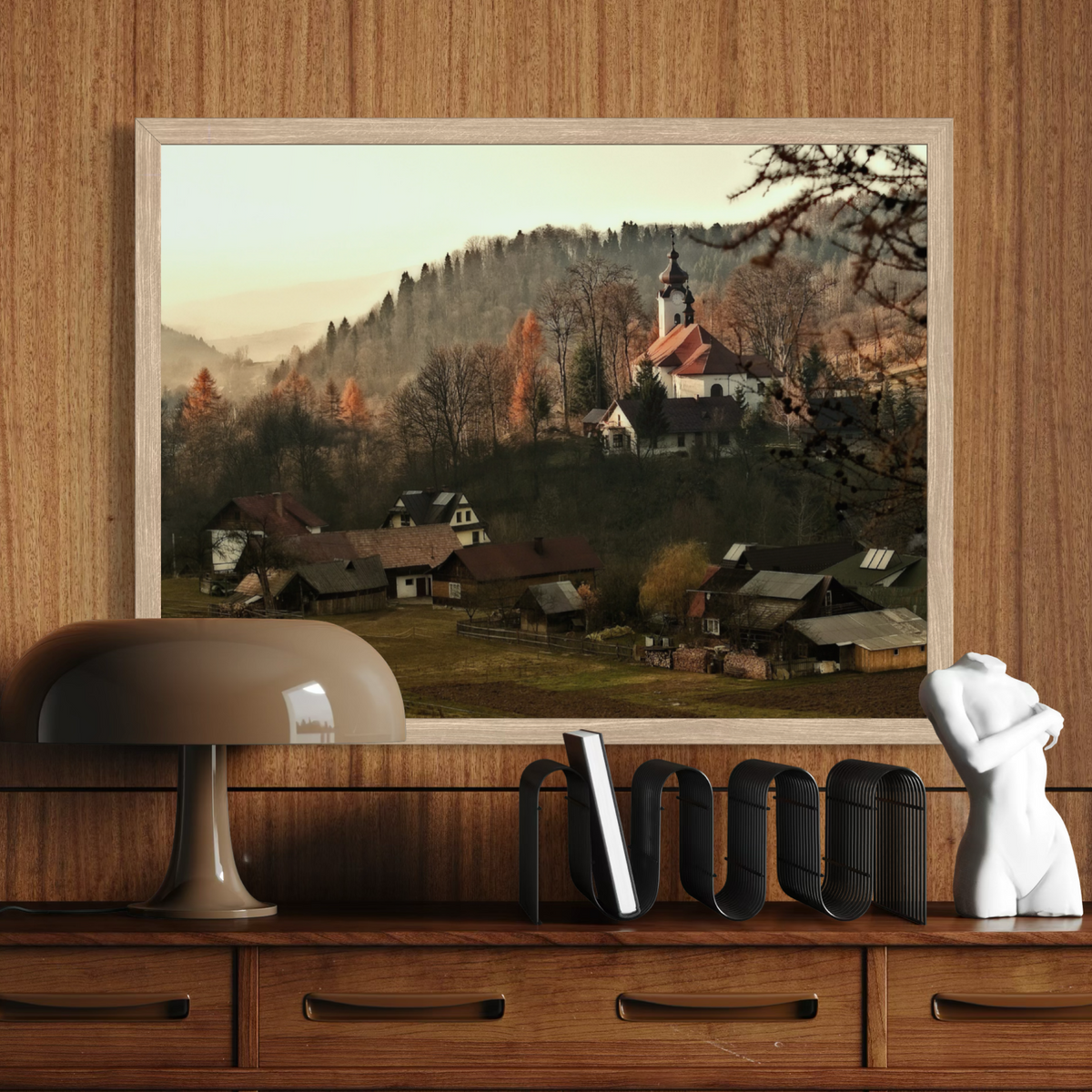 A framed landscape art print depicting a quaint village with a church on a hill, displayed on a wooden shelf next to a lamp and a white bust sculpture.