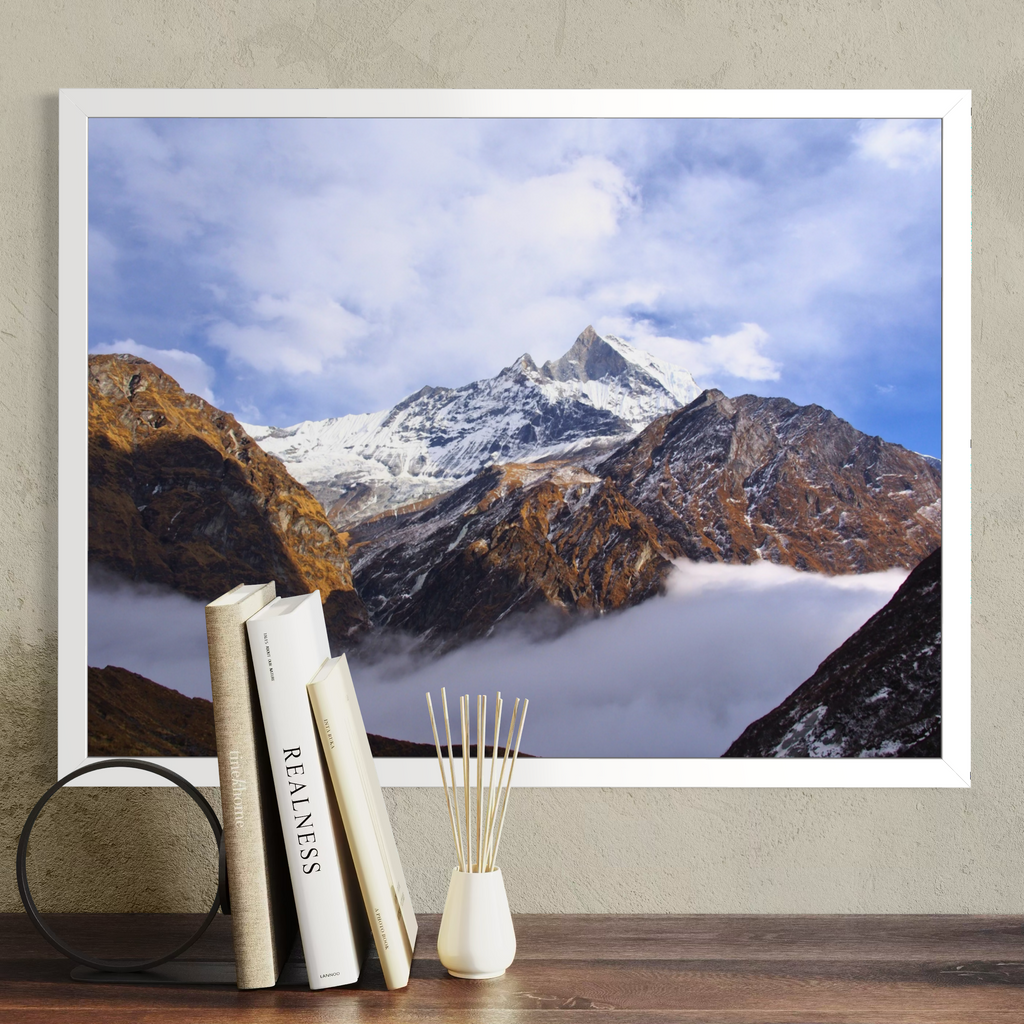 A large framed wall art print showing a snow-capped mountain peak above clouds, displayed on a beige wall above a wooden surface with books, a black circular decor object, and a small white vase with reeds.