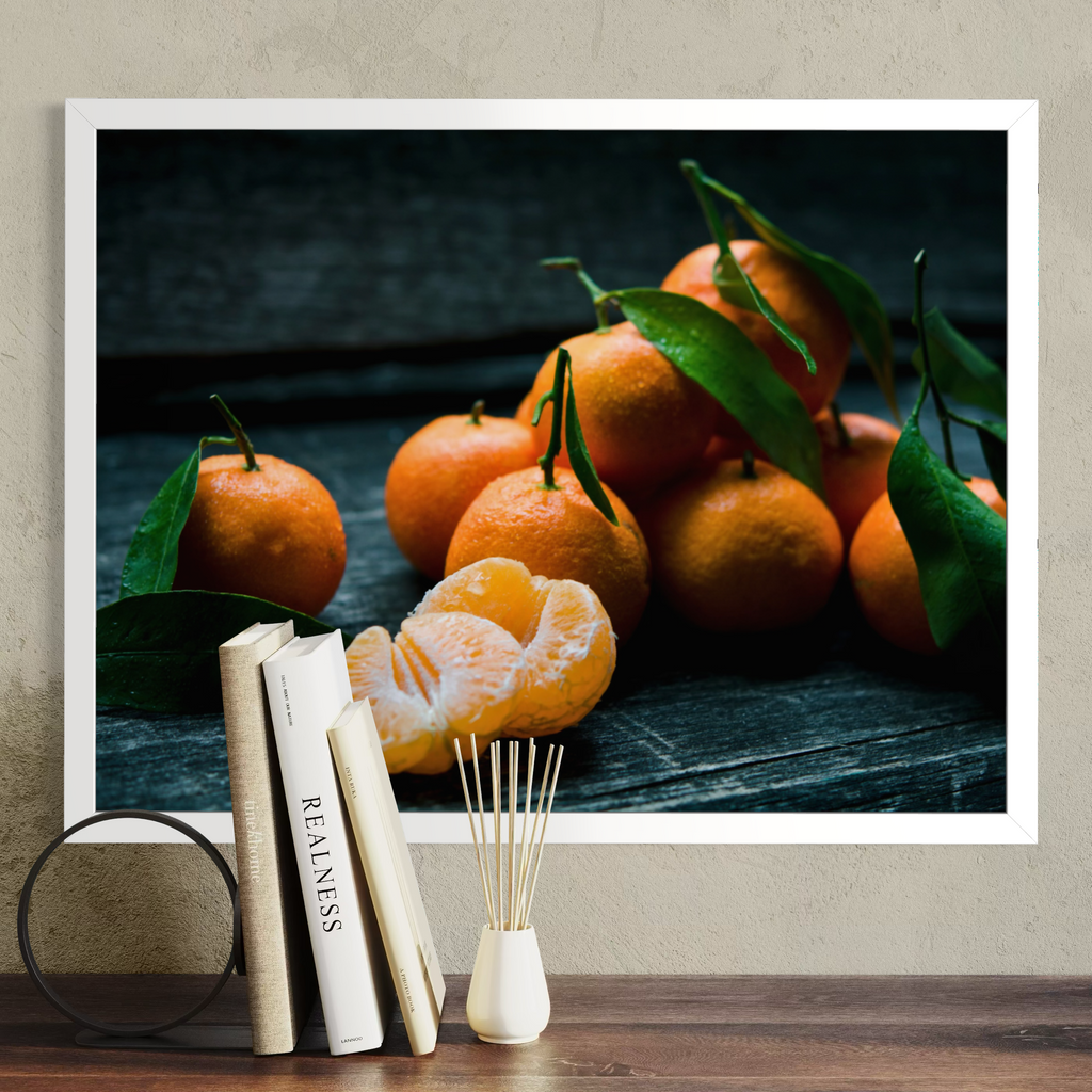 A framed wall art print showing a still life of oranges with green leaves on a dark wooden surface, displayed above a tabletop with books and a reed diffuser against a textured beige wall.