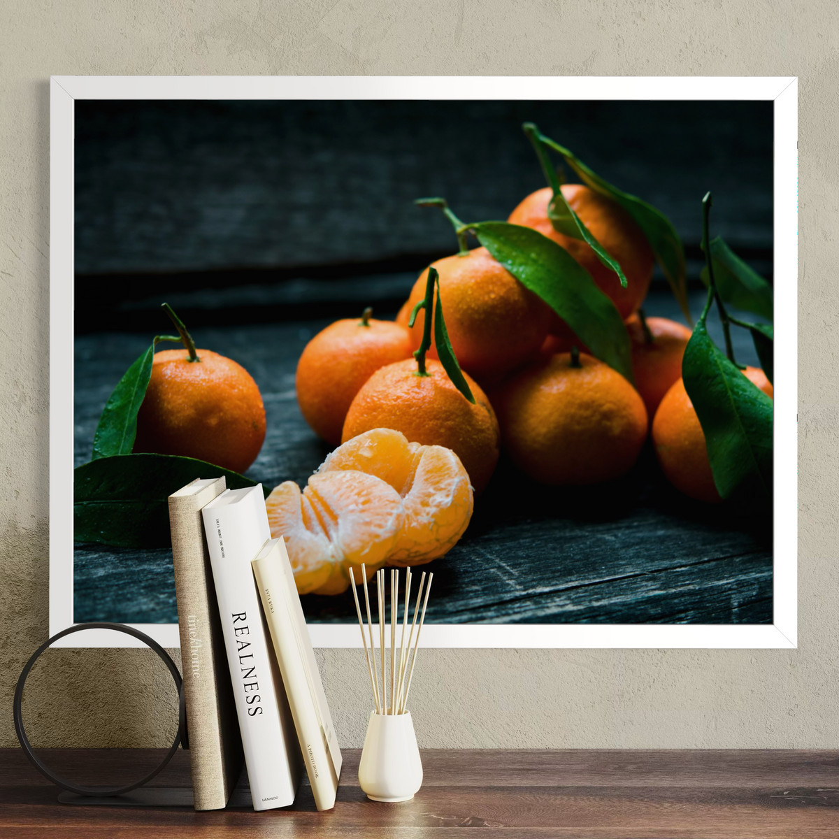 A framed wall art print showing a still life of oranges with green leaves on a dark wooden surface, displayed above a tabletop with books and a reed diffuser against a textured beige wall.