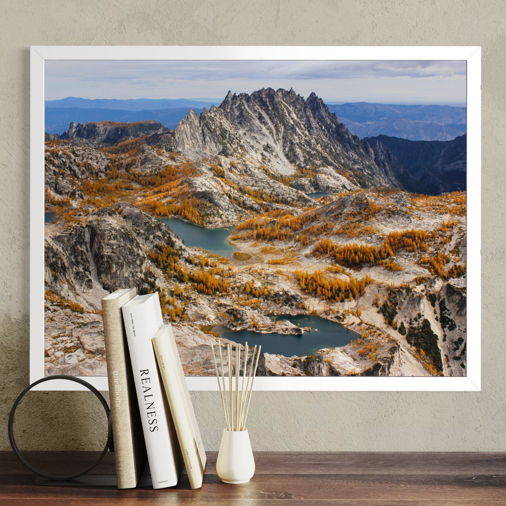 A white framed landscape art print of a mountain range with blue lakes and golden autumn trees, displayed on a wall above a wooden table with books and a reed diffuser.