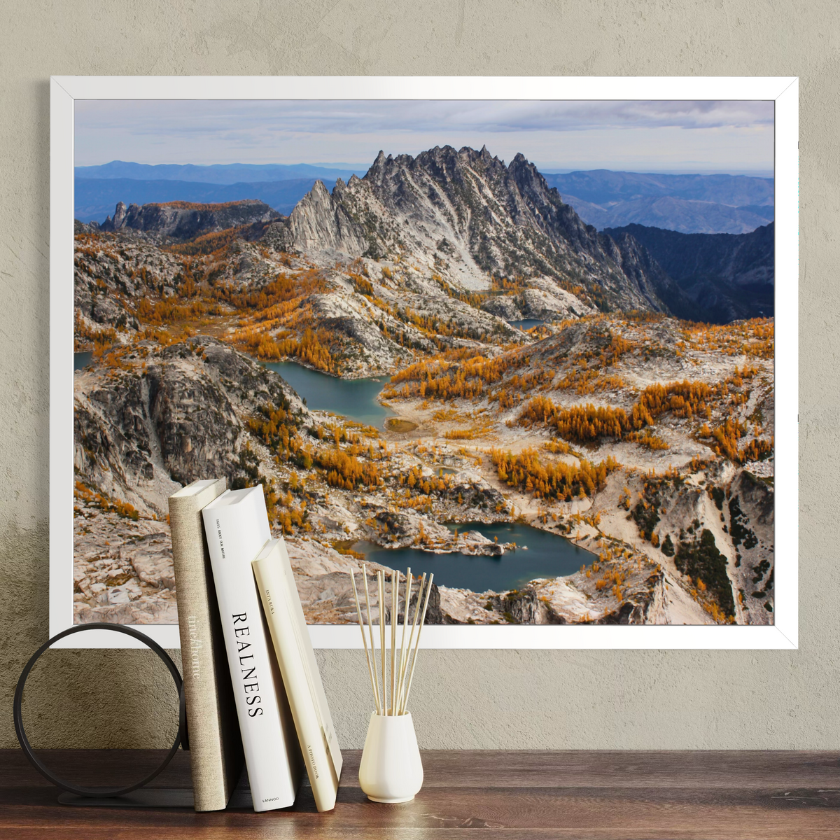 A white framed landscape art print of a mountain range with blue lakes and golden autumn trees, displayed on a wall above a wooden table with books and a reed diffuser.