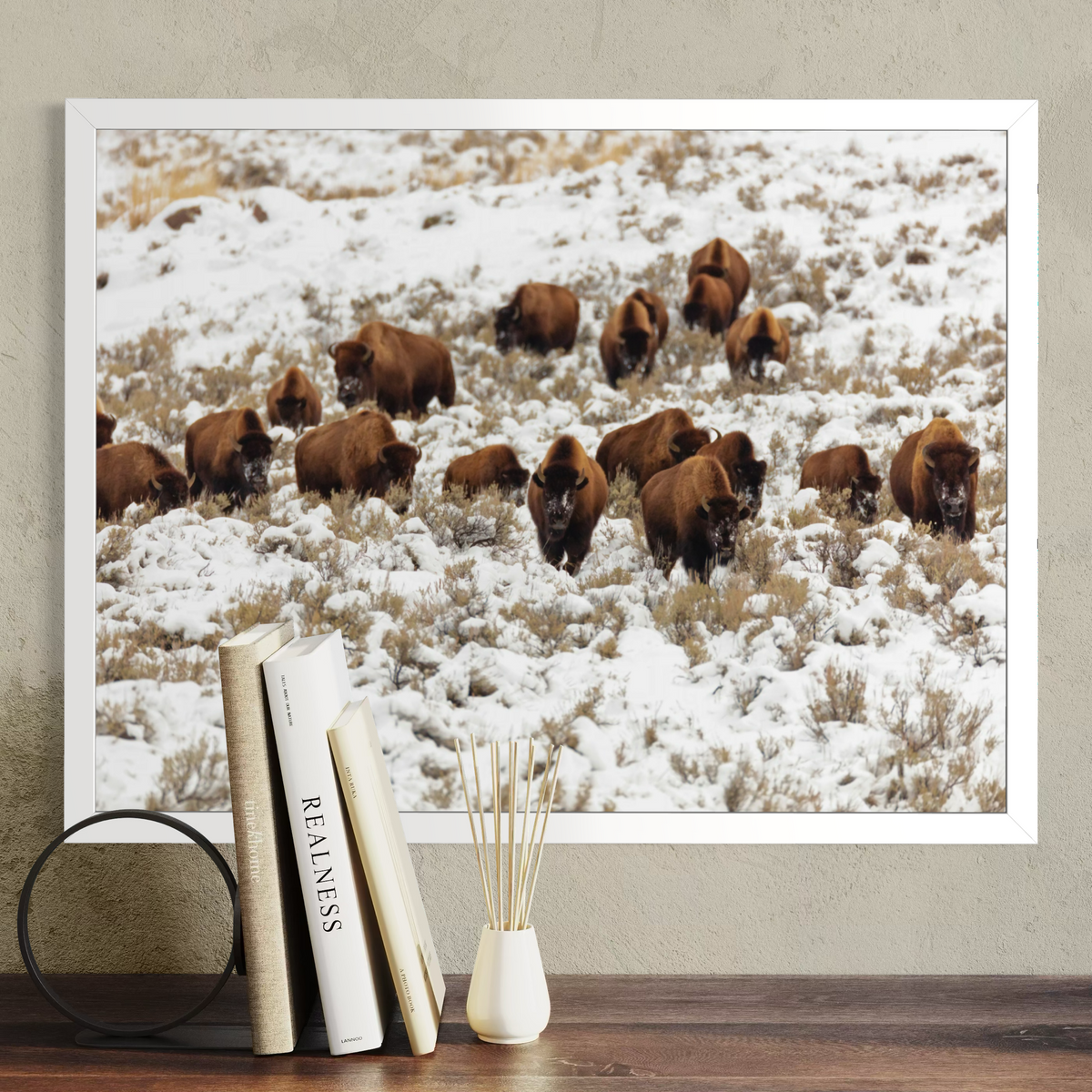 A white framed wall art print showing a herd of brown bison standing in a snowy field, displayed above a wooden surface with books and a small vase against a textured beige wall.
