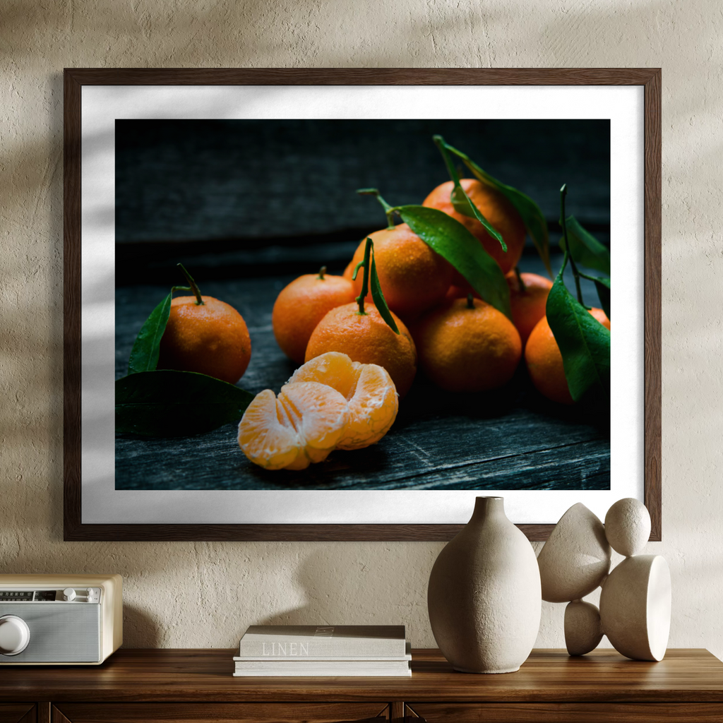 A framed wall art print featuring a still life of orange clementines with green leaves on a dark rustic surface, displayed in a warm beige room above a wooden console table with decorative objects.