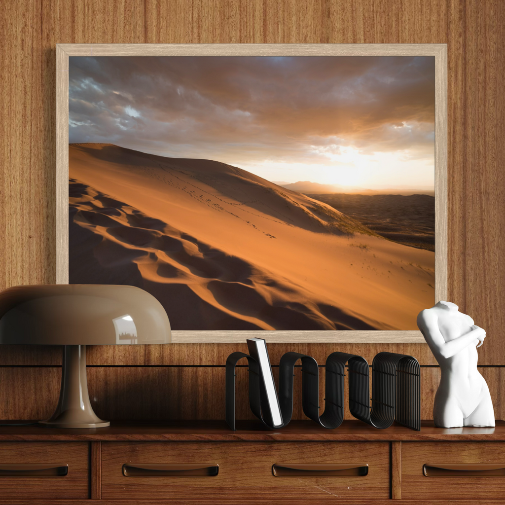 A framed wall art print depicting golden desert sand dunes under a dramatic sunset sky, displayed on a wooden wall above a console table with a lamp, books, and a white torso sculpture.