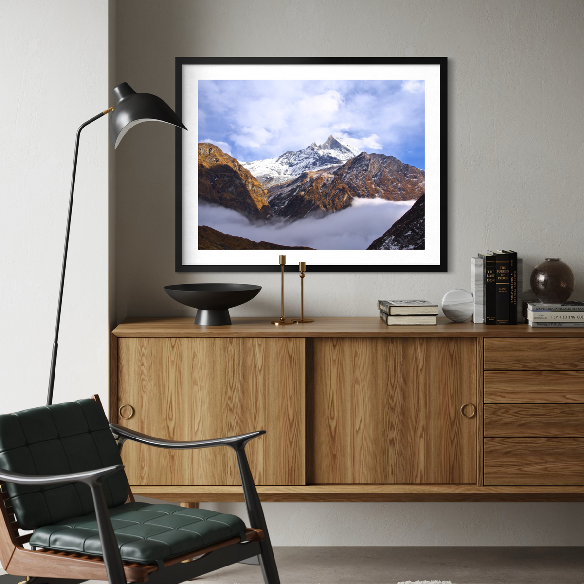 A framed landscape art print of snow-capped mountains under a blue sky, displayed on a light gray wall above a wooden credenza in a modern living room setting.