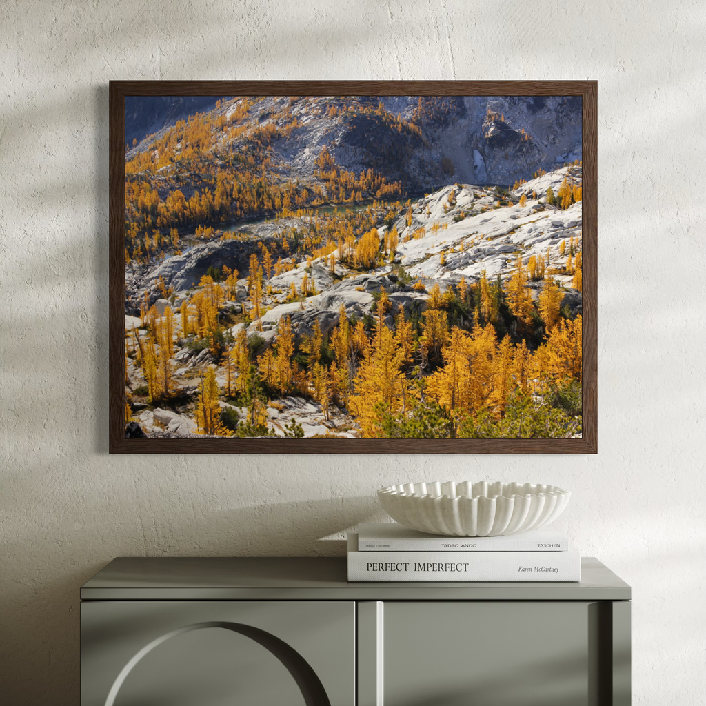 A wooden framed landscape art print depicting a mountain range with golden larchwood trees and snow, displayed on a textured white wall above a gray console table with books and a decorative bowl.