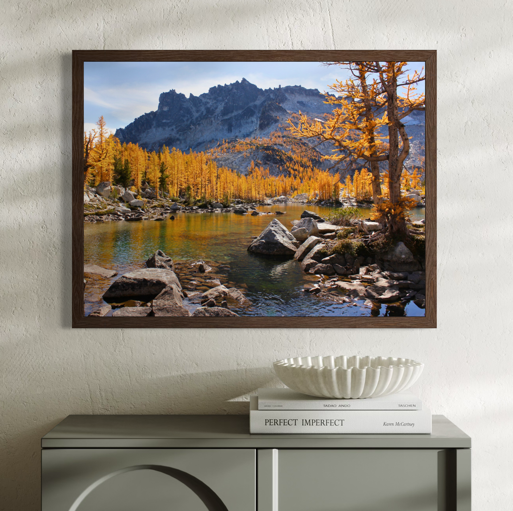 A wooden framed landscape art print depicting a mountain lake with golden larchwood trees, displayed on a textured white wall above a gray console table with a white decorative bowl and books.