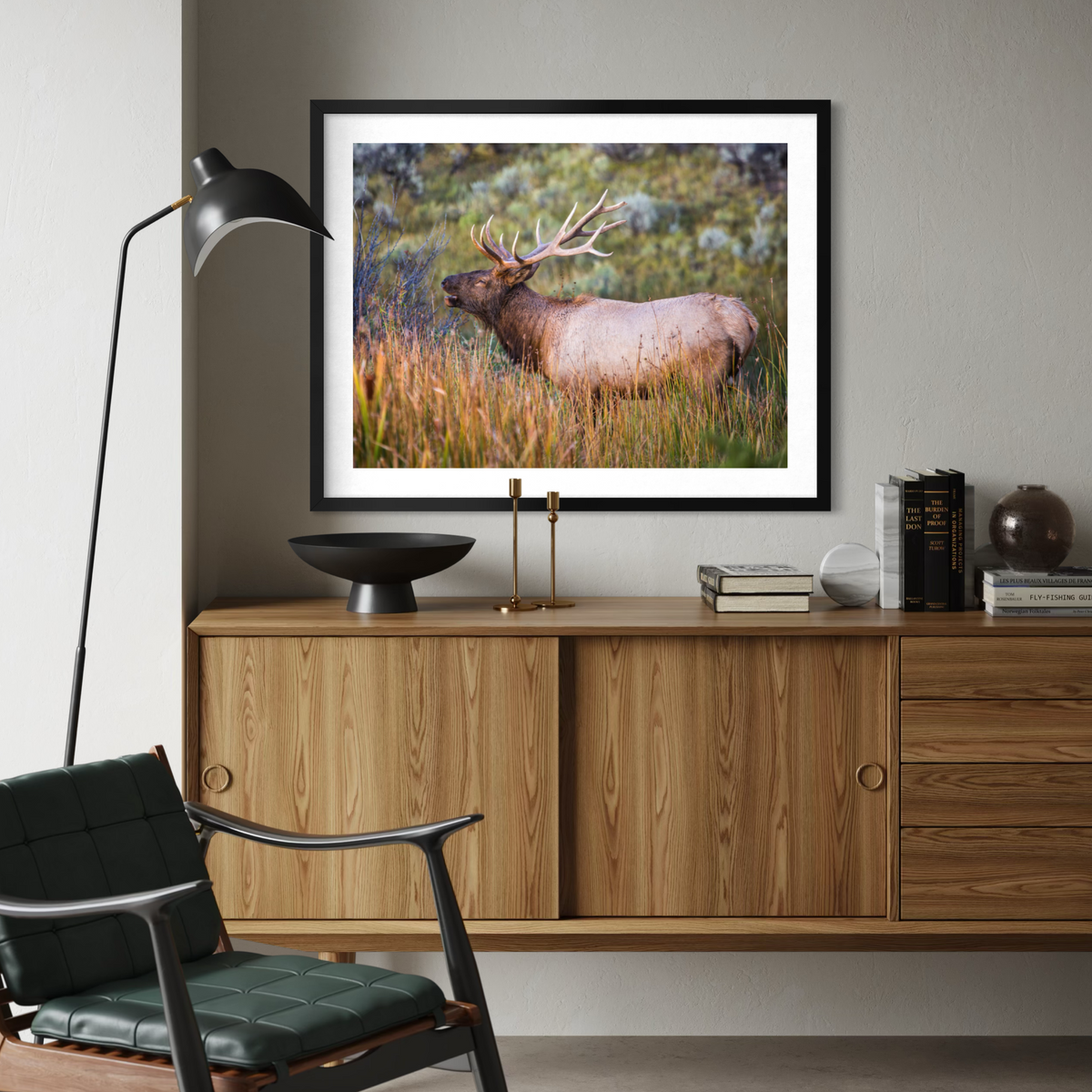 A black framed art print of an elk in a grassy field, displayed on a light gray wall above a mid-century modern wooden credenza in a styled living room setting.