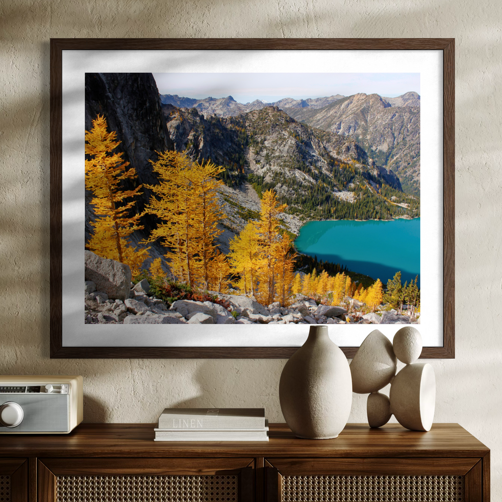 A wooden framed landscape art print featuring golden larchets, rocky mountains, and a turquoise lake, displayed on a wooden console table against a textured beige wall.