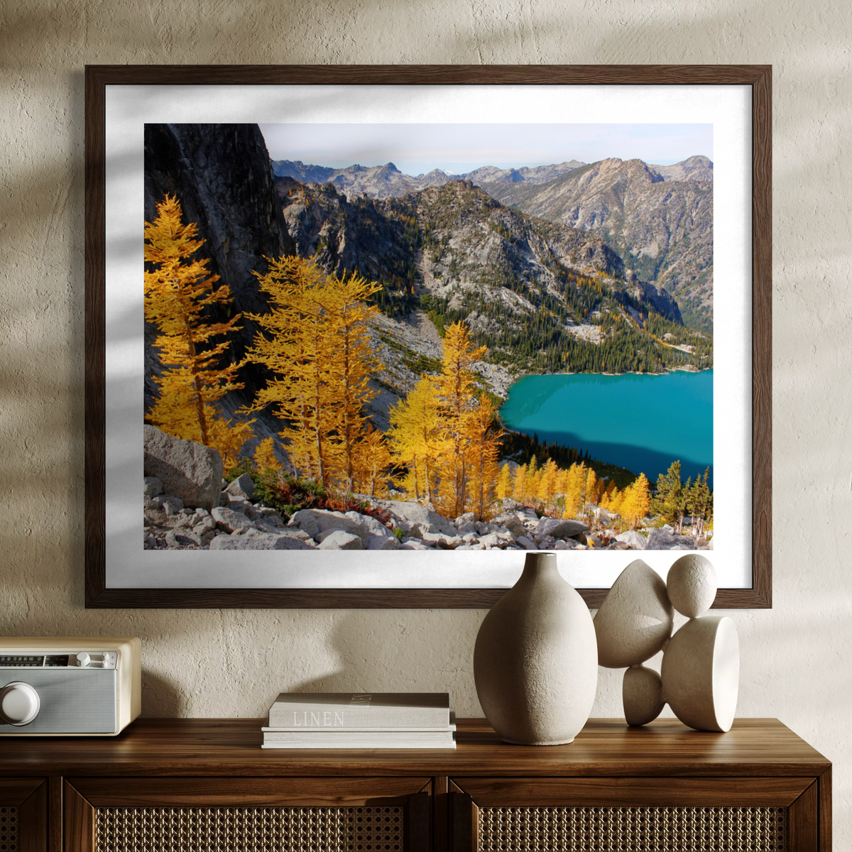 A wooden framed landscape art print featuring golden larchets, rocky mountains, and a turquoise lake, displayed on a wooden console table against a textured beige wall.