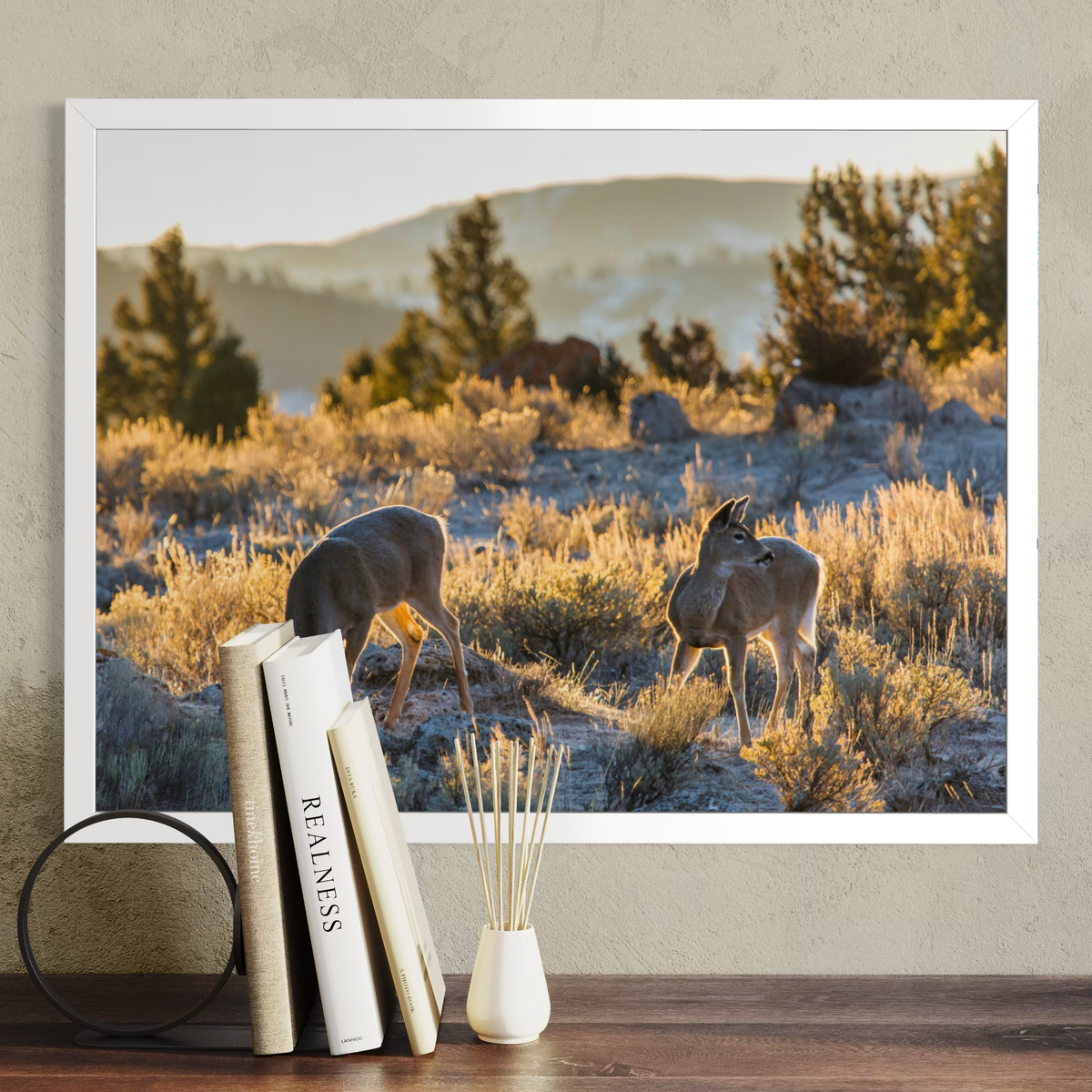 A framed landscape art print showing two deer in a grassy field at sunset, displayed on a wall above a wooden table with books and a small vase.