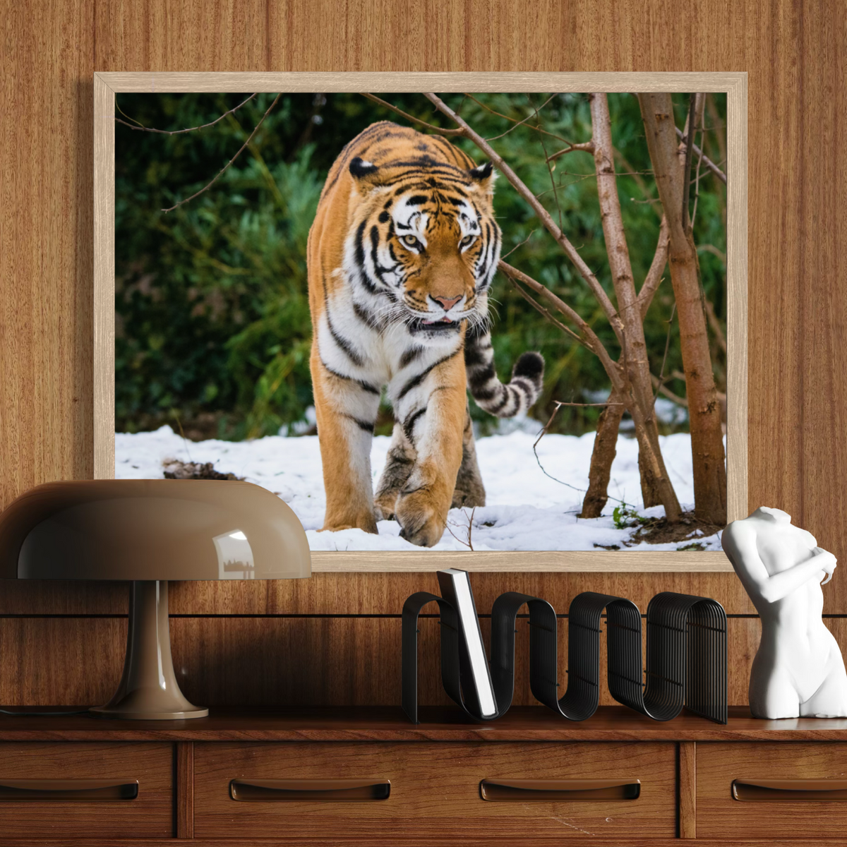 A wooden framed wall art print depicting a tiger walking through snow with trees in the background, mounted on a wood-paneled wall above a wooden console table with a lamp, bookends, and a small white sculpture.
