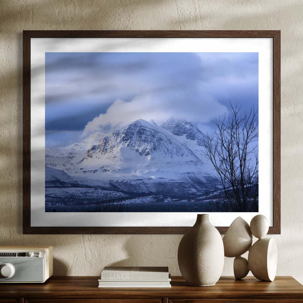 A framed landscape art print of a snow-covered mountain under a cloudy blue sky, displayed on a textured beige wall above a wooden console table with decorative vases and a radio.
