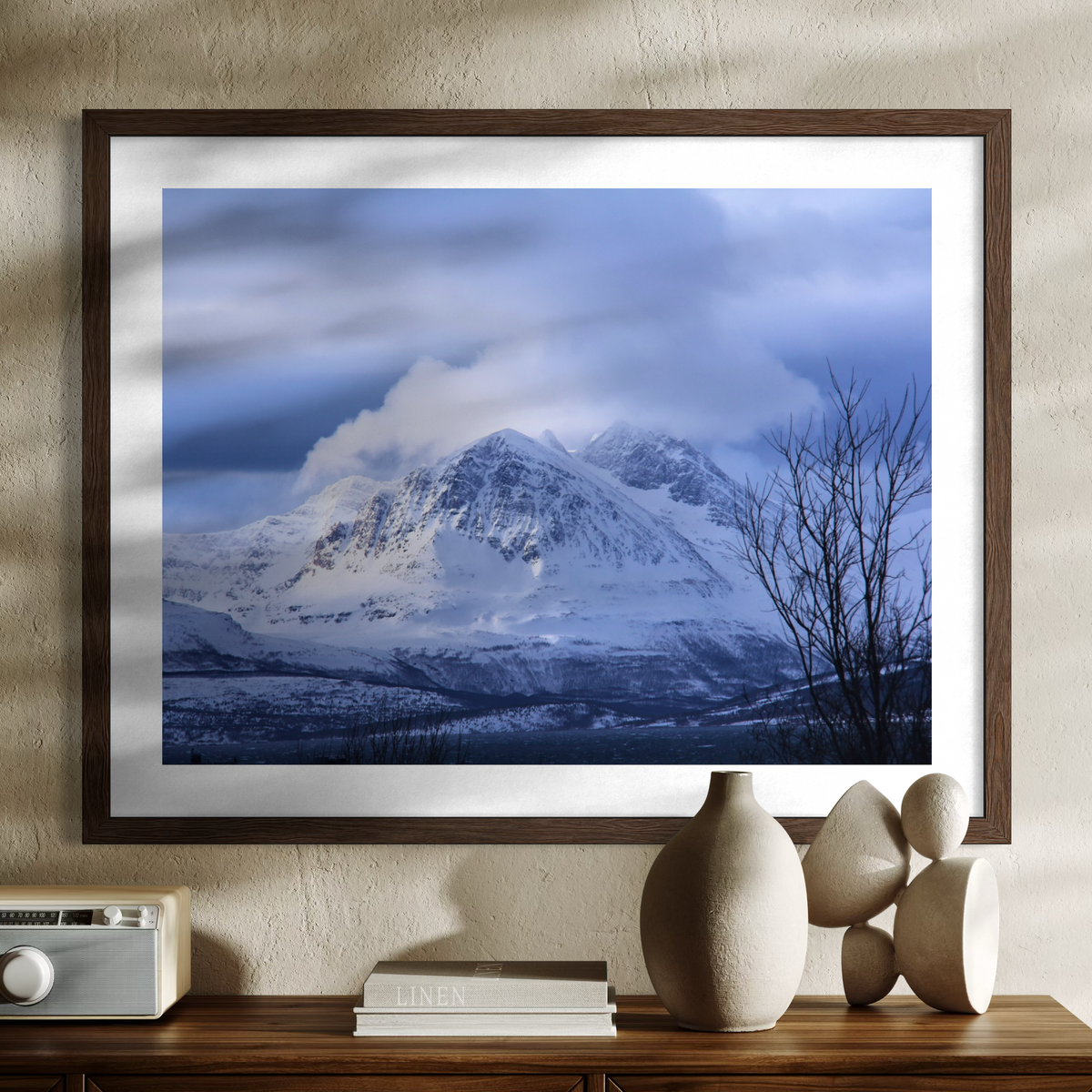 A framed landscape art print of a snow-covered mountain under a cloudy blue sky, displayed on a textured beige wall above a wooden console table with decorative vases and a radio.