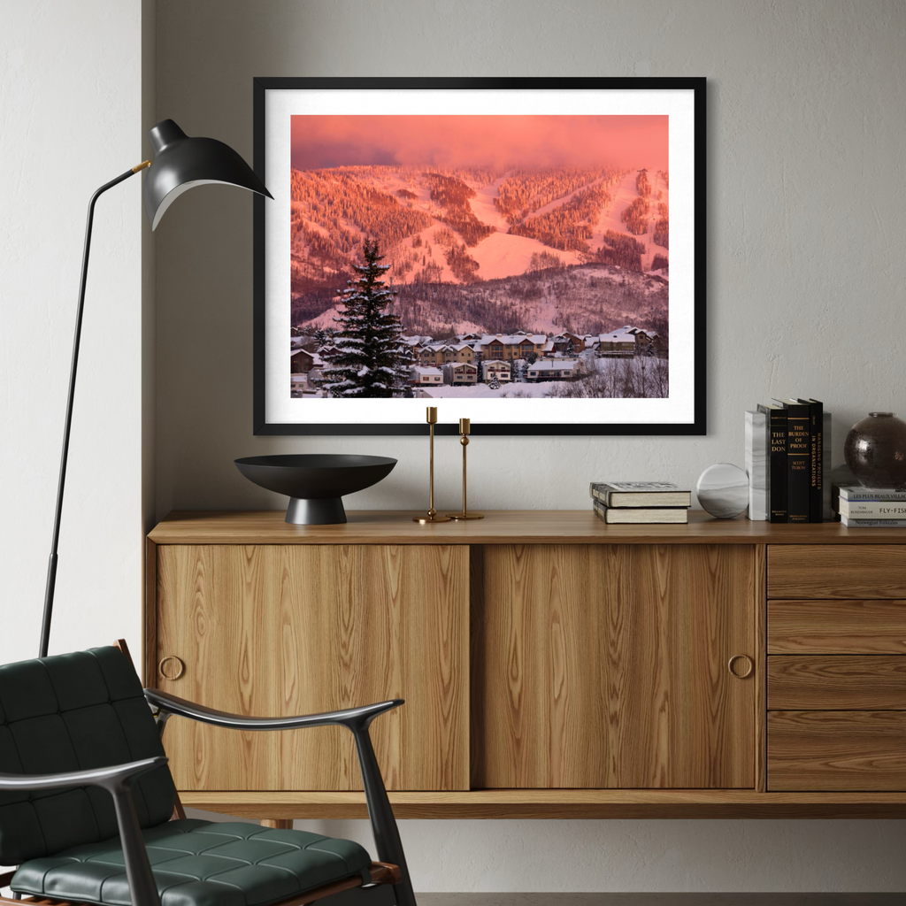 A framed art print depicting a snowy mountain village under a pink and orange sunset sky, styled on a light gray wall above a wooden credenza in a modern living room setting.