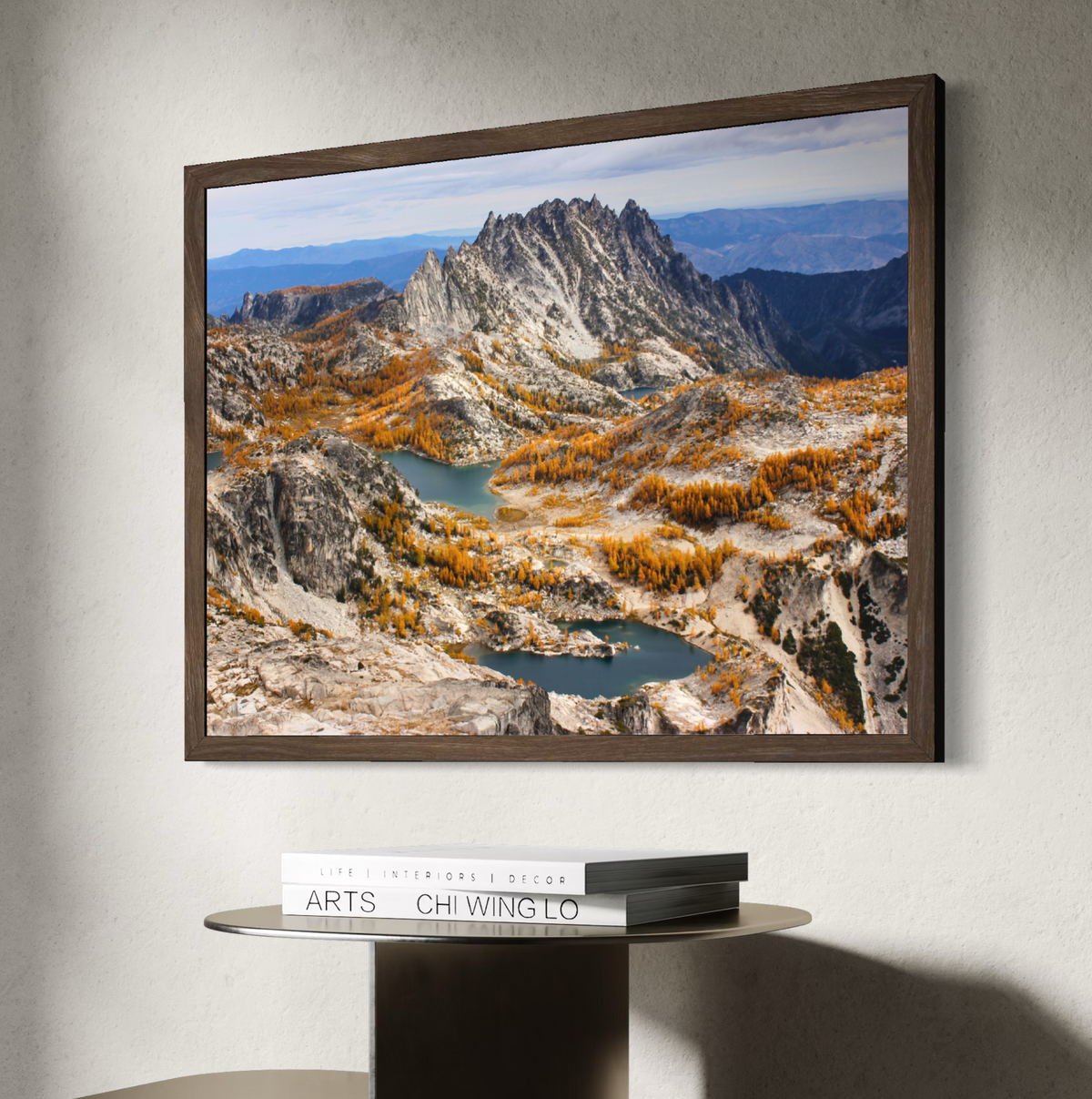 A wooden framed landscape art print depicting a mountain range with blue lakes and golden autumn trees, displayed on a wall above a side table with books.
