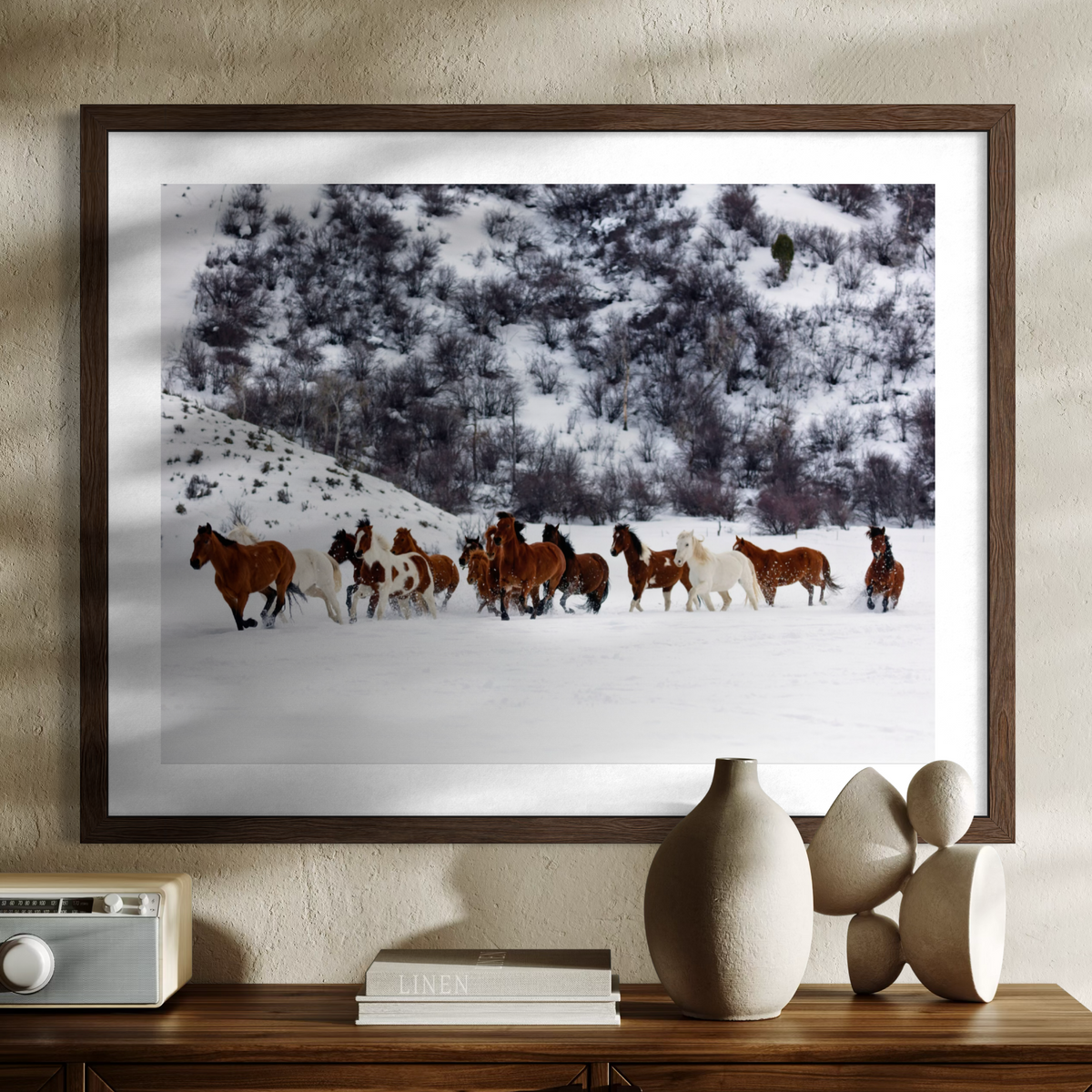 A framed art print depicting a herd of horses running through a snowy mountain landscape, displayed on a textured beige wall above a wooden console table with decorative vases and books.