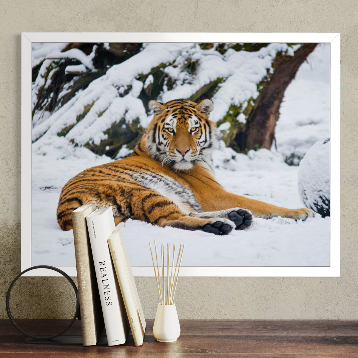 A white framed wall art print showing a tiger lying in a snowy outdoor scene, displayed on a beige wall above a wooden surface with books and a small vase in front.