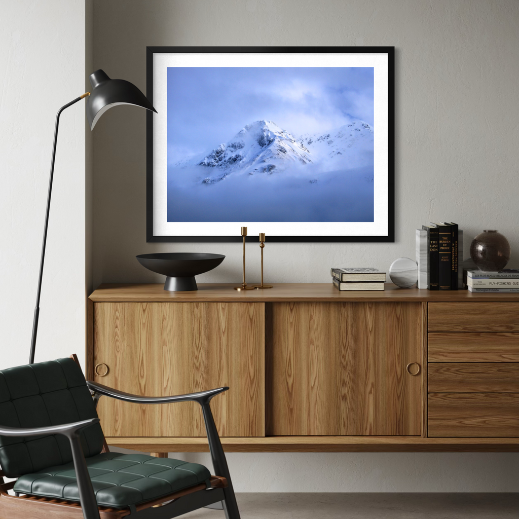 A framed art print of a snowy mountain peak under a blue sky, displayed on a wooden console table in a modern living room setting.