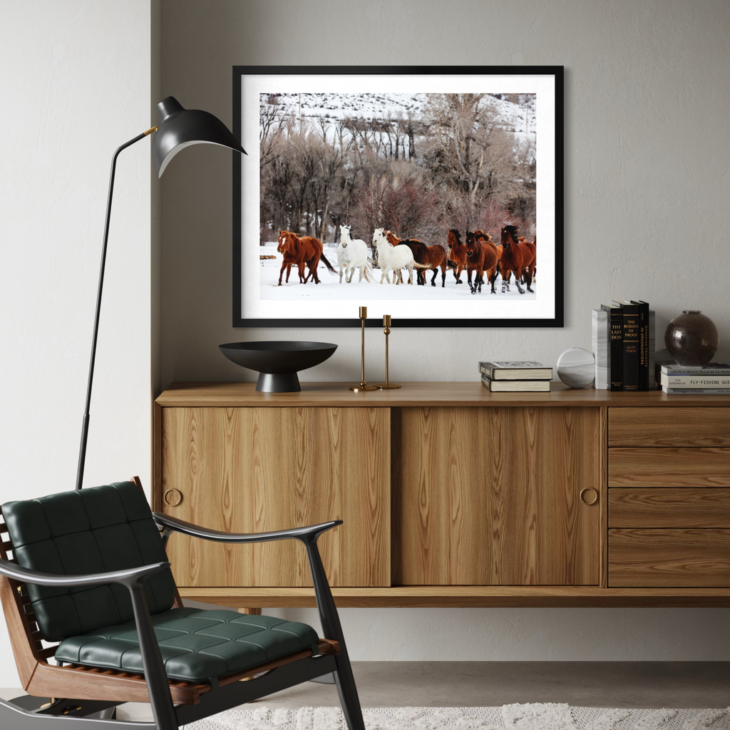 A framed art print depicting a herd of horses in a snowy landscape, displayed on a wooden sideboard in a modern living room setting.