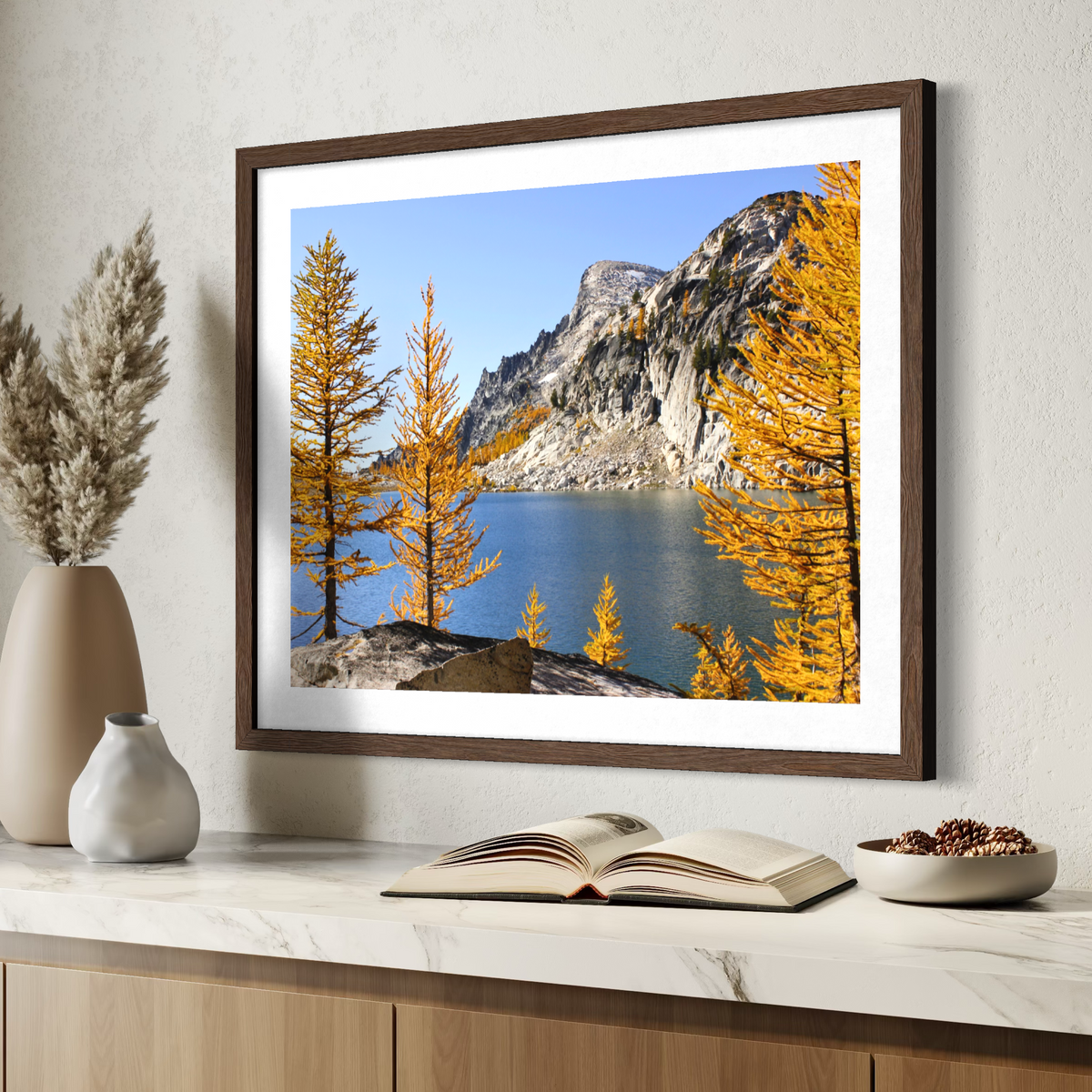 A wooden framed landscape art print depicting a mountain lake with golden yellow trees under a blue sky, styled on a white marble countertop with decorative vases, an open book, and a bowl of pinecones.