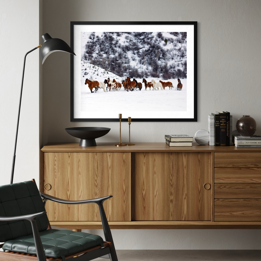 A framed art print depicting a herd of horses in a snowy landscape, displayed on a light gray wall above a mid-century modern wooden credenza in a styled living room setting.