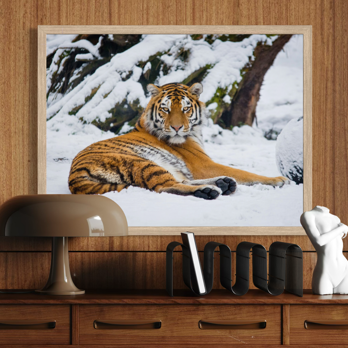 A wooden framed wall art print depicting a tiger lying in snow, displayed above a wooden console table with a lamp, books, and a small white sculpture in a styled interior setting.