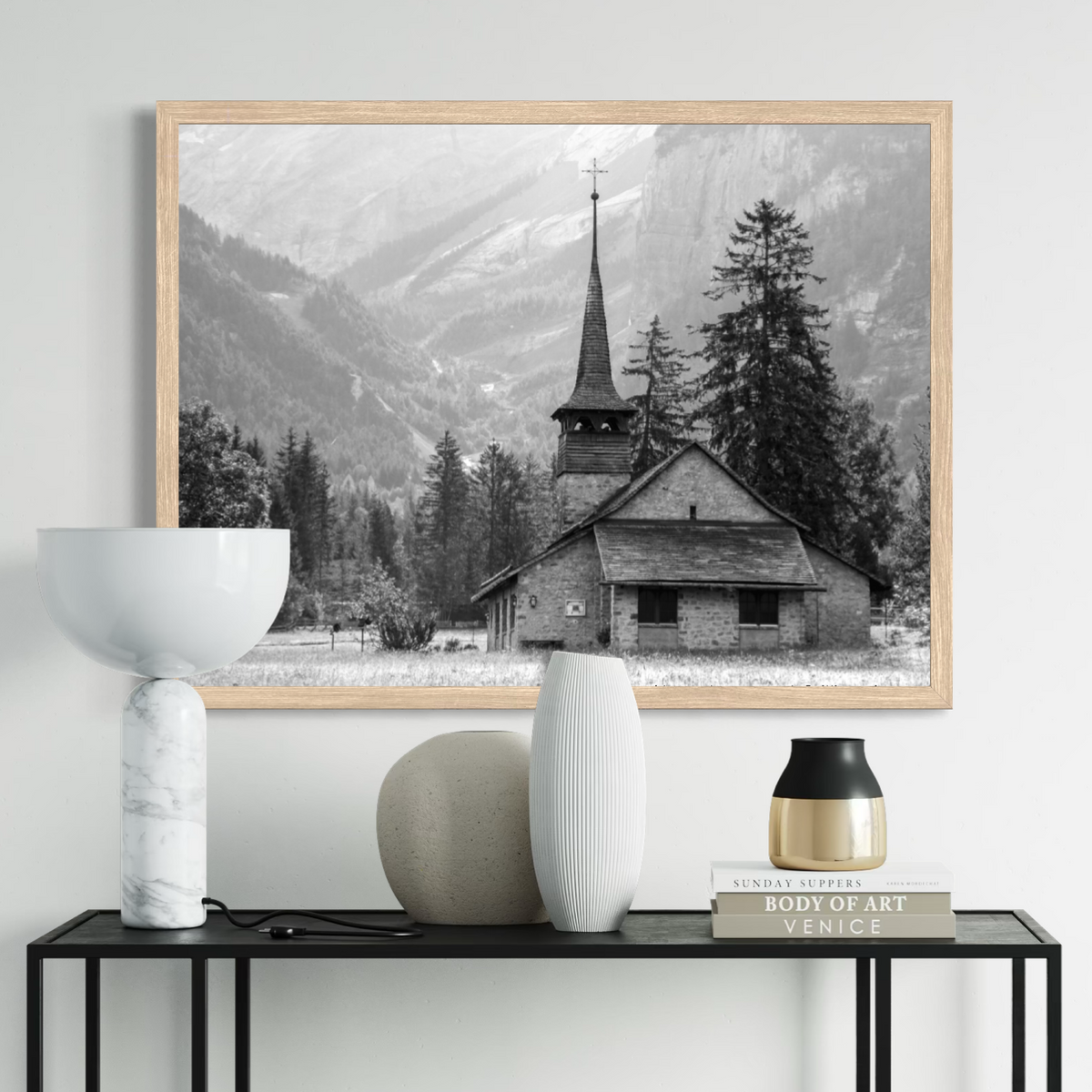 A framed black and white landscape art print of a mountain village church, displayed on a light gray wall above a console table with decorative vases and books.