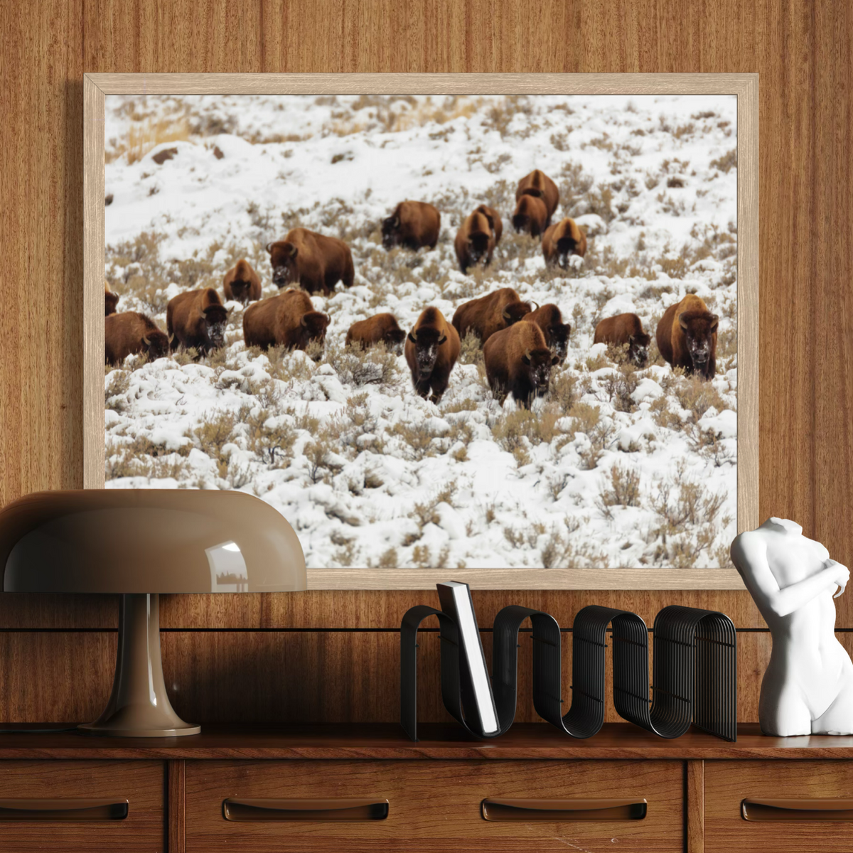 A framed wall art print depicting a herd of brown bison in a snowy field, displayed on a wooden console above a lamp and books.
