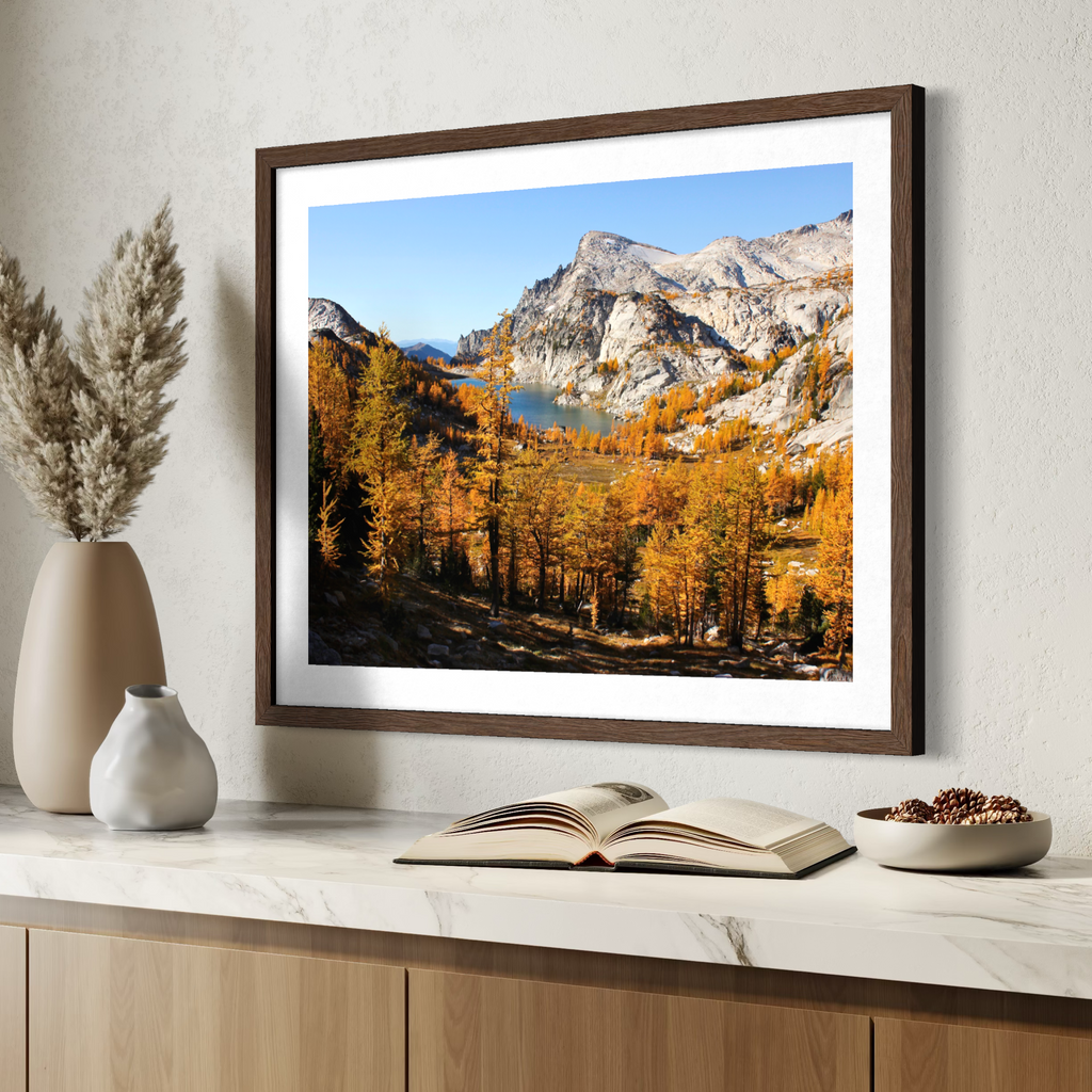 A wooden framed landscape art print depicting autumn mountains, a lake, and golden trees, styled on a marble countertop with decorative vases, an open book, and a bowl of seeds.