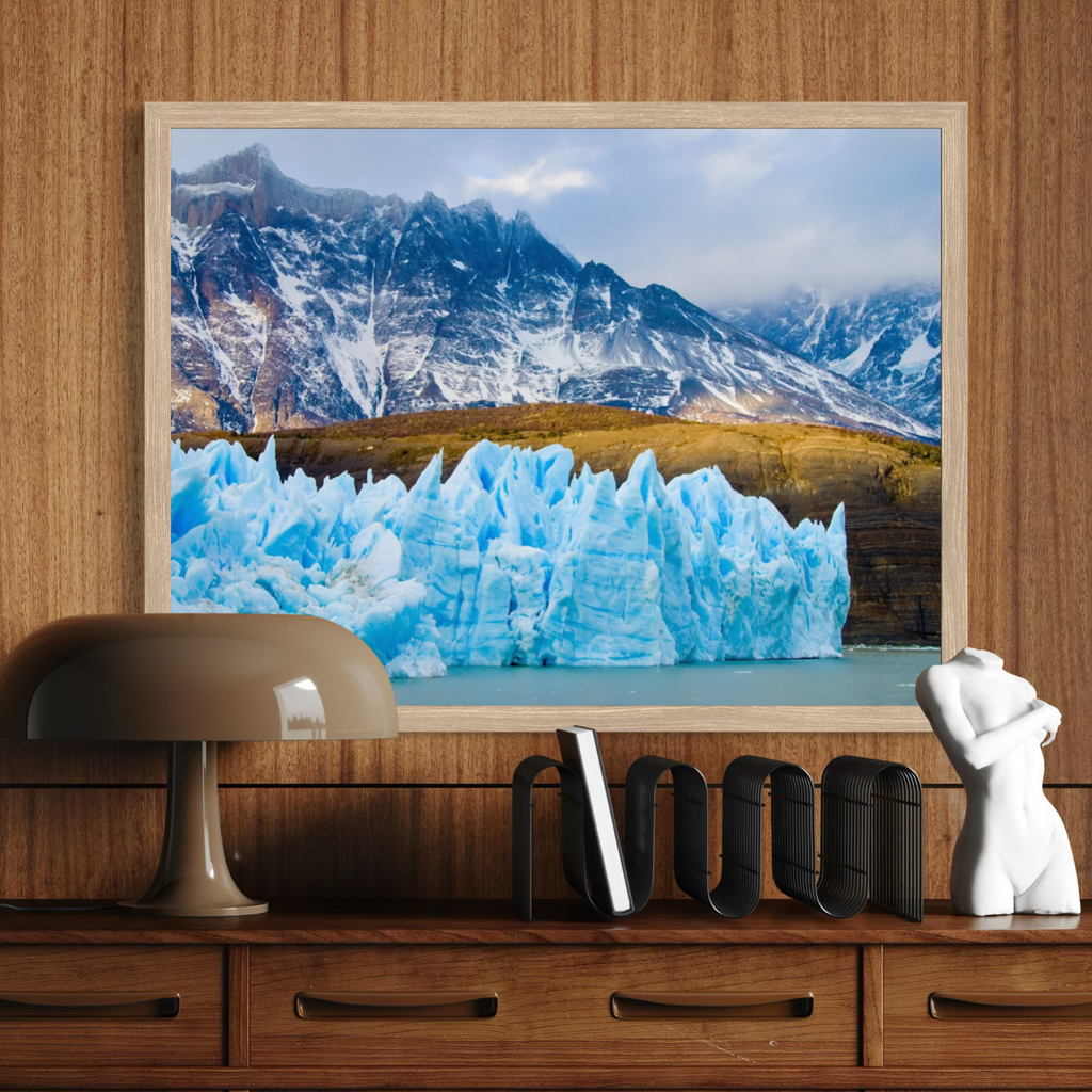 A framed landscape art print depicting a glacier and snowy mountains, styled on a wooden console table with a lamp, books, and a small white sculpture.
