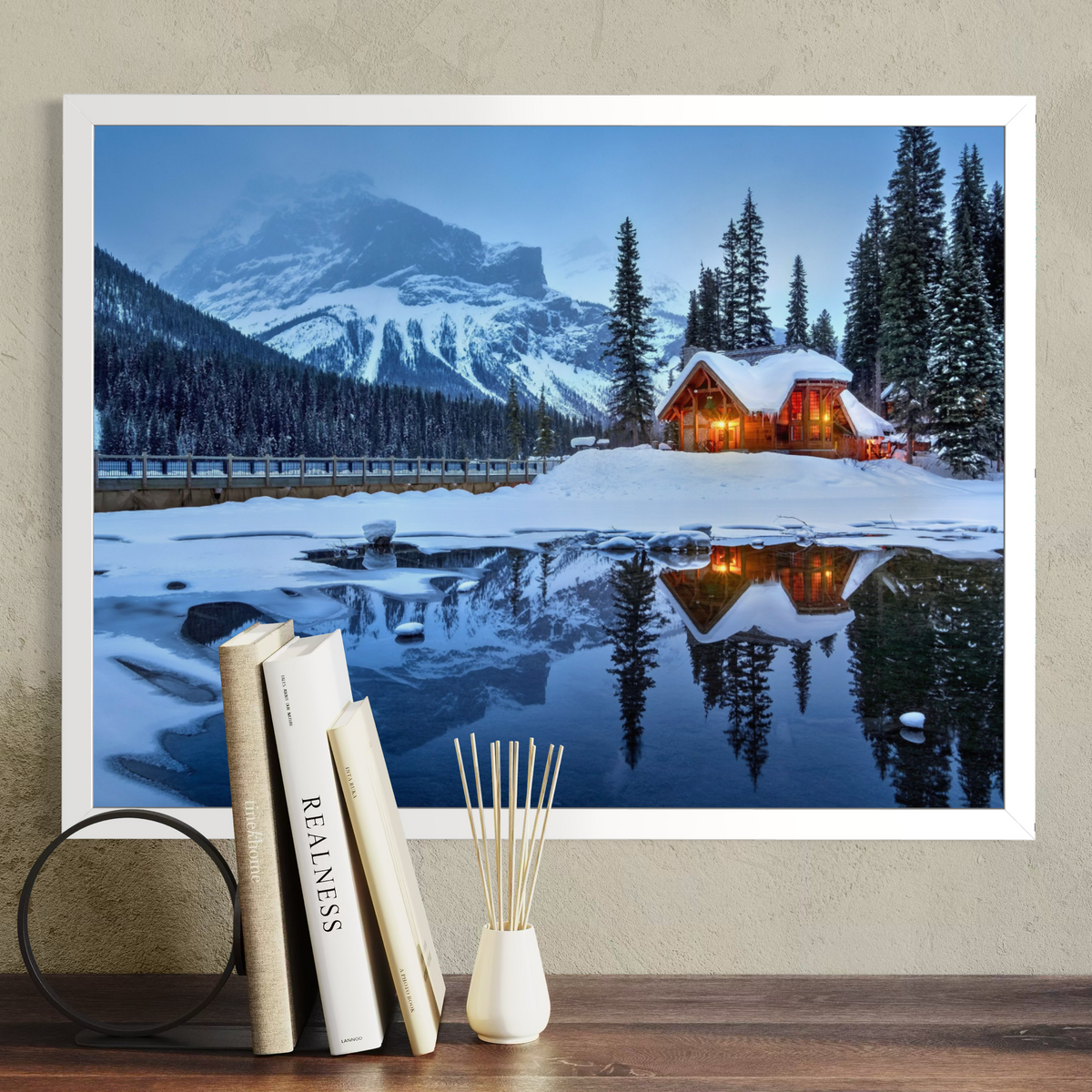 A framed wall art print showing a snowy mountain landscape with a glowing cabin reflected in a calm lake, displayed above a wooden surface with books and a reed diffuser in a styled interior setting.