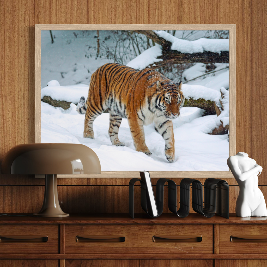 A wooden framed wall art print showing a tiger walking through a snowy outdoor scene, displayed above a wooden console table with a lamp, books, and a small white figurine.