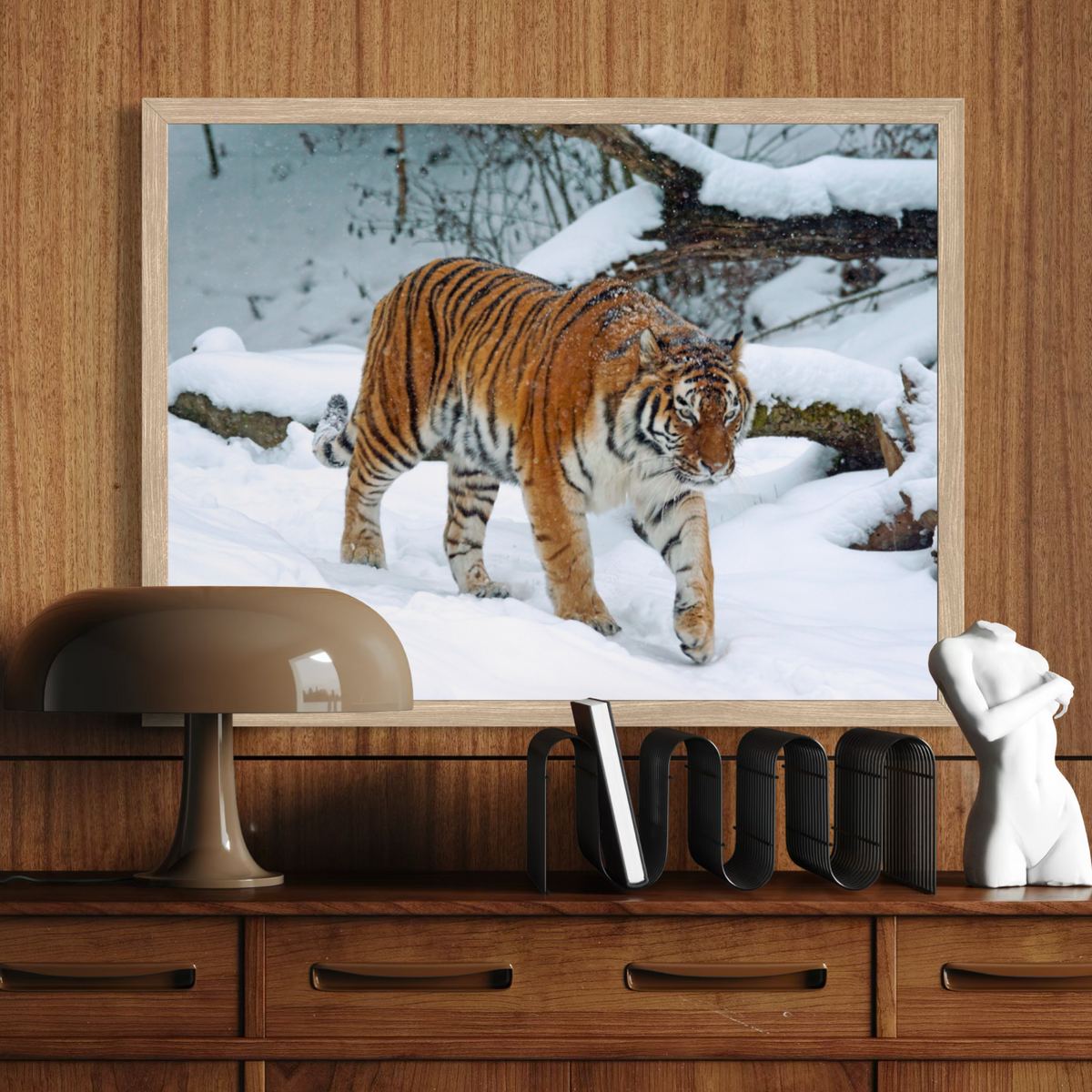 A wooden framed wall art print showing a tiger walking through a snowy outdoor scene, displayed above a wooden console table with a lamp, books, and a small white figurine.