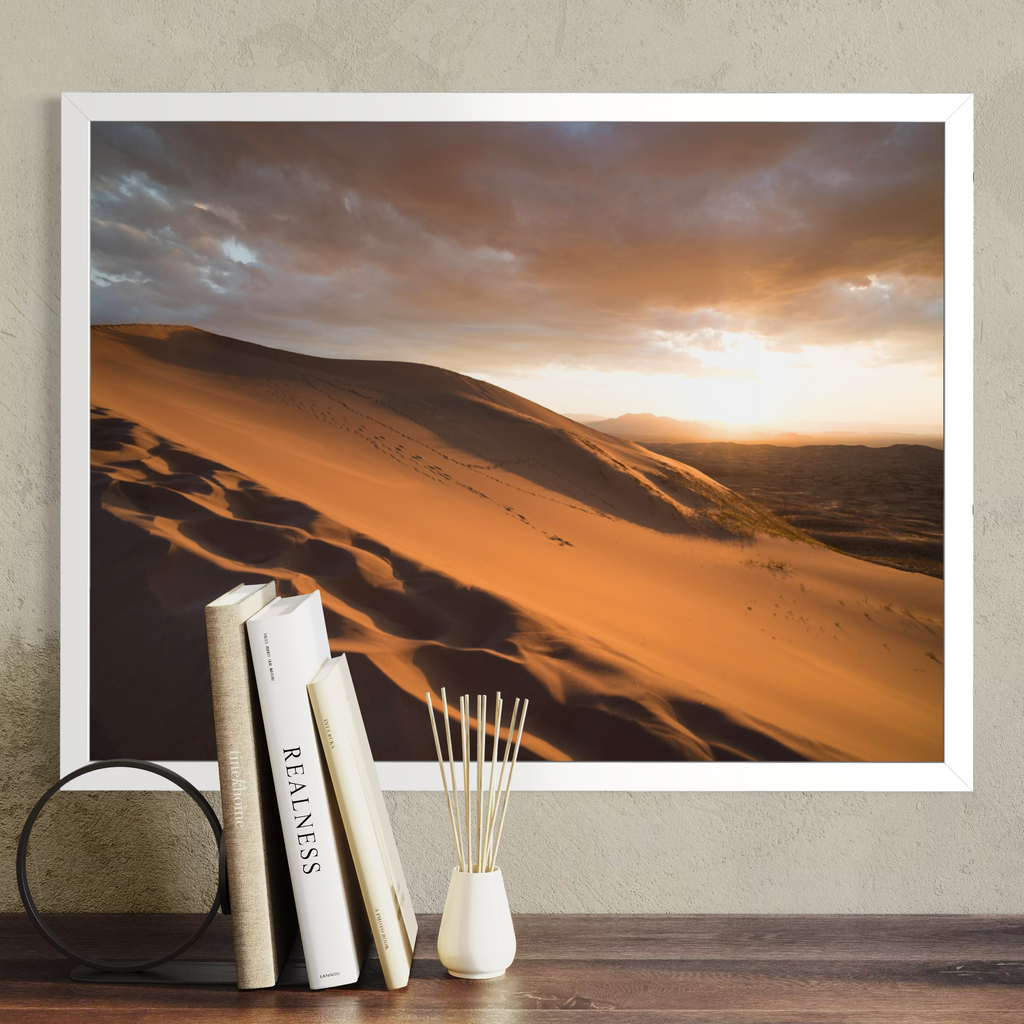 A framed desert landscape print hangs on a textured beige wall above a wooden surface, styled with three books and a small white vase with reeds. The print shows golden sand dunes under a dramatic cloudy sky at sunset.