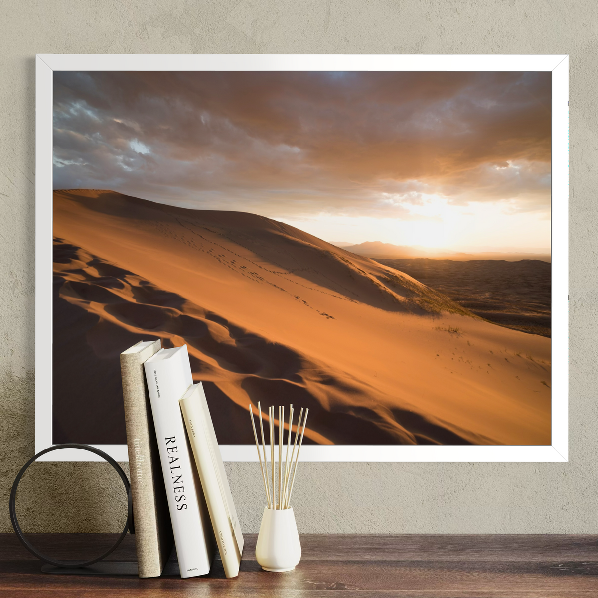 A framed desert landscape print hangs on a textured beige wall above a wooden surface, styled with three books and a small white vase with reeds. The print shows golden sand dunes under a dramatic cloudy sky at sunset.