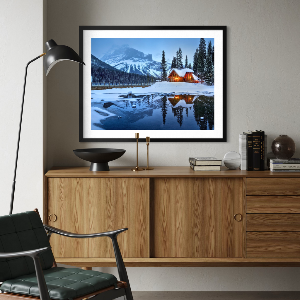 A framed landscape art print depicting a snowy mountain lake with glowing cabin lights, displayed on a wall above a wooden console table in a modern living room setting.