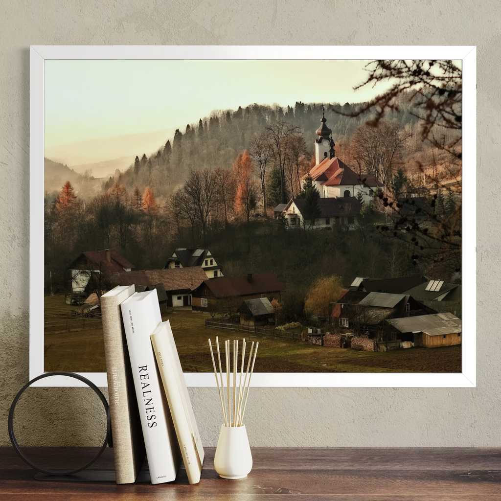 A framed landscape art print depicting a quaint village with a church on a hill, displayed on a shelf next to books and a reed diffuser against a textured wall.