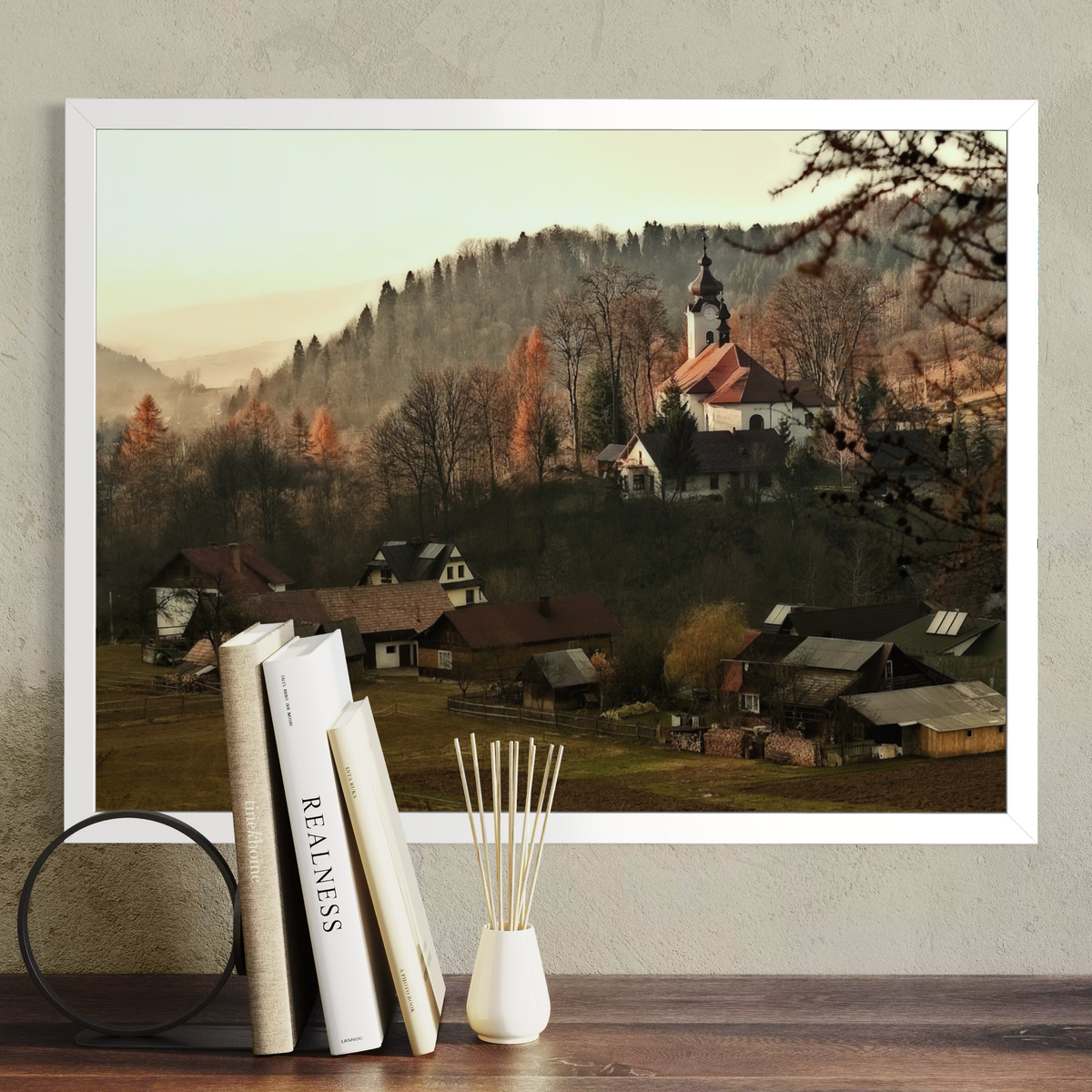 A framed landscape art print depicting a quaint village with a church on a hill, displayed on a shelf next to books and a reed diffuser against a textured wall.