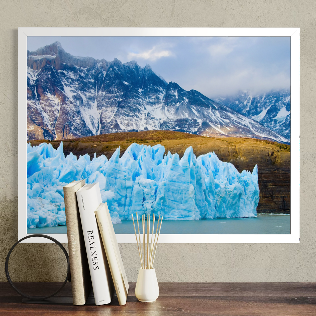 A framed landscape art print of a glacier and snowy mountains, styled on a wooden table with books and a small vase against a textured beige wall.