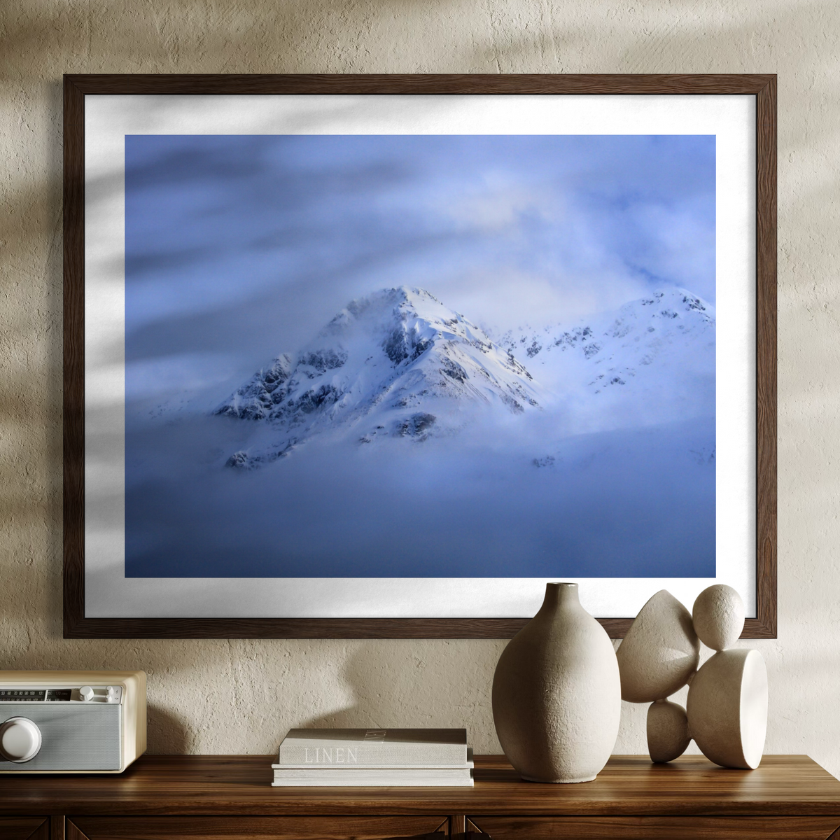 A framed art print depicting a snowy mountain peak shrouded in blue clouds, hung on a textured beige wall above a wooden console table with decorative ceramic vases and books.