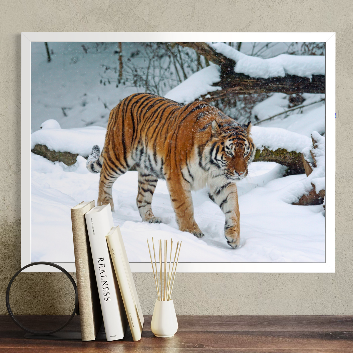 A white framed photographic print of a tiger walking through a snowy forest, displayed on a beige wall above a wooden surface with books and a reed diffuser in front.