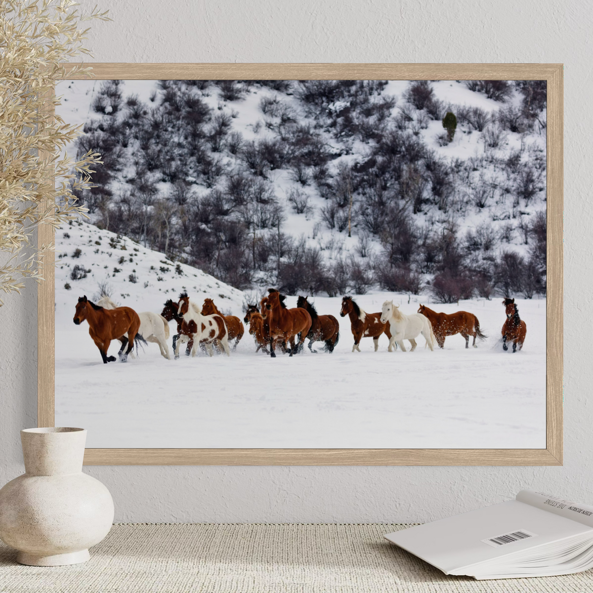 A framed art print depicting a herd of wild horses running through a snowy mountain landscape, styled on a tabletop next to a ceramic vase and an open book.