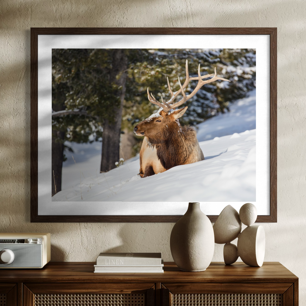 A wooden framed wall art print showing a large deer with antlers resting in a snowy forest, displayed above a wooden console table with decorative vases and books against a textured beige wall.