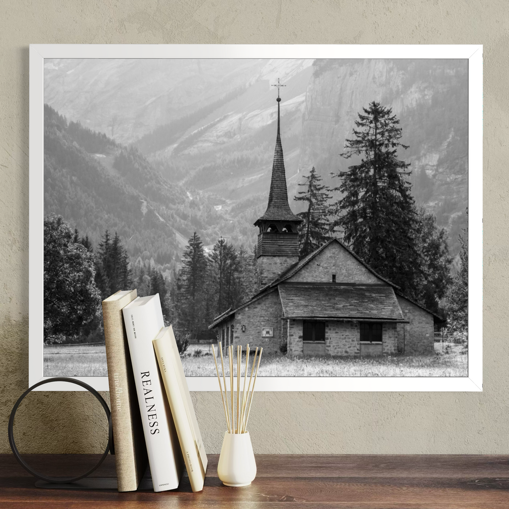 A framed black and white landscape photograph of a church with a tall steeple, set against mountains and evergreen trees, displayed on a beige wall above a wooden table with books and a small vase.